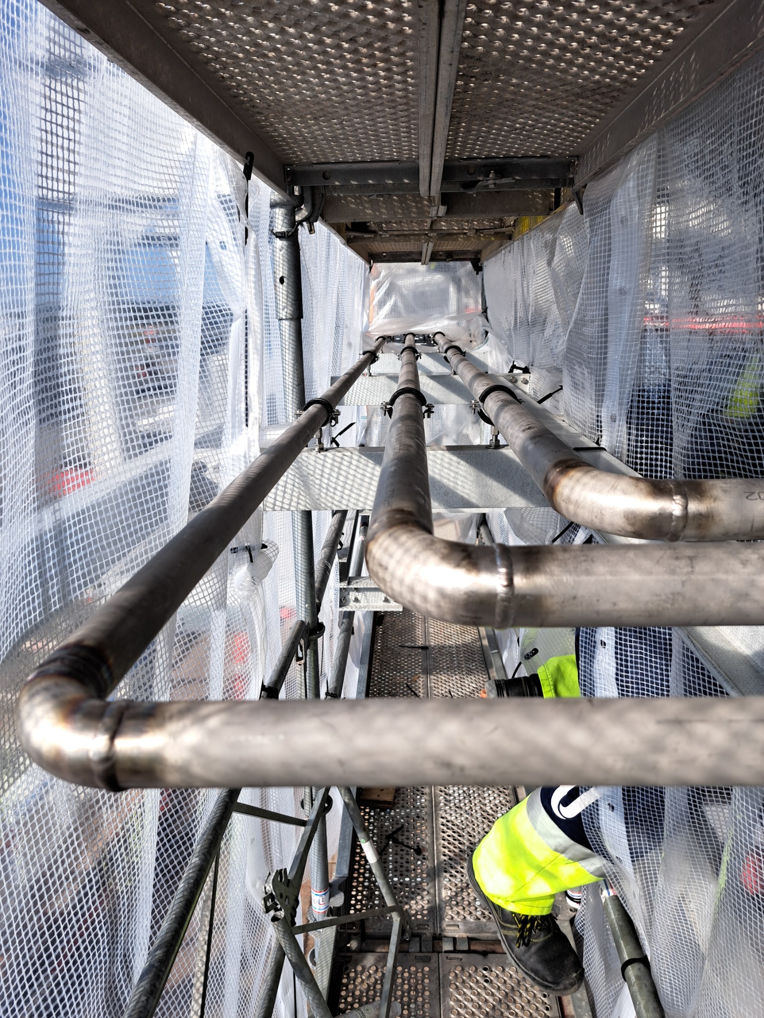 Spojrzenie z góry na spawane rury na rusztowaniu, zabezpieczone siatką ochronną. Widoczne złącza spawane i fragment stroju roboczego.