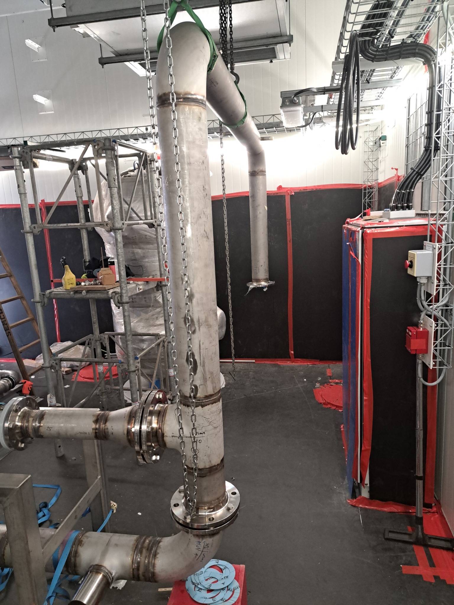Instalacja rur przemysłowych ze stali nierdzewnej, spawanych i łączonych kołnierzami, zawieszona na łańcuchach w hali produkcyjnej. Widoczne rusztowania i elementy instalacji elektrycznej.