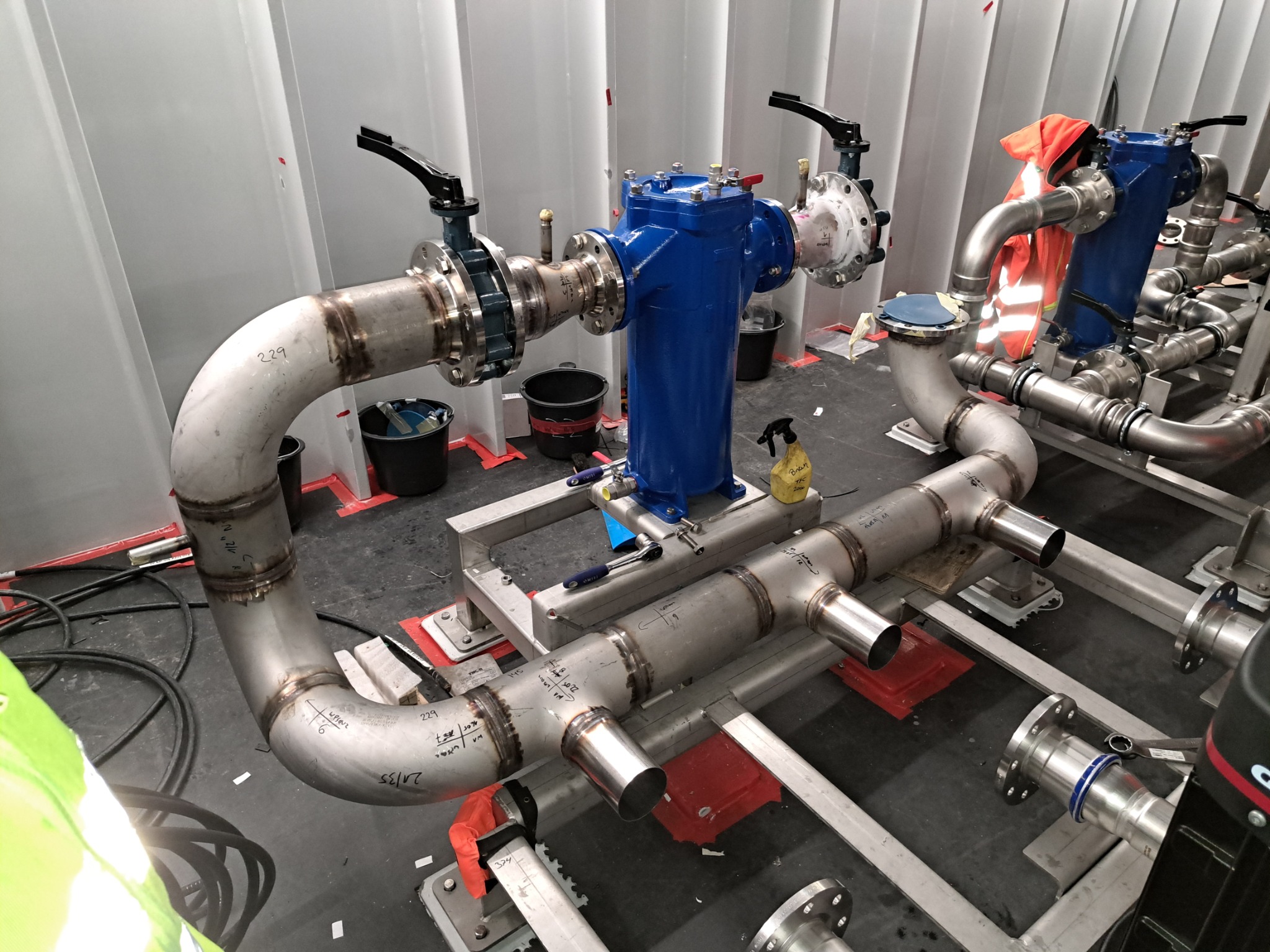 Złożona instalacja rurociągowa ze stali nierdzewnej z niebieskim filtrem i zaworami, po spawaniu, z widocznymi oznaczeniami i narzędziami w hali.