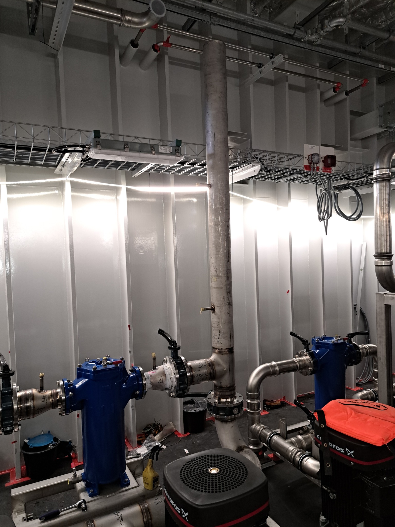 Instalacja przemysłowa z rurami i zaworami w hali, widoczne elementy spawane. Białe ściany i oświetlenie. Elementy instalacji w kolorze srebrnym i niebieskim.