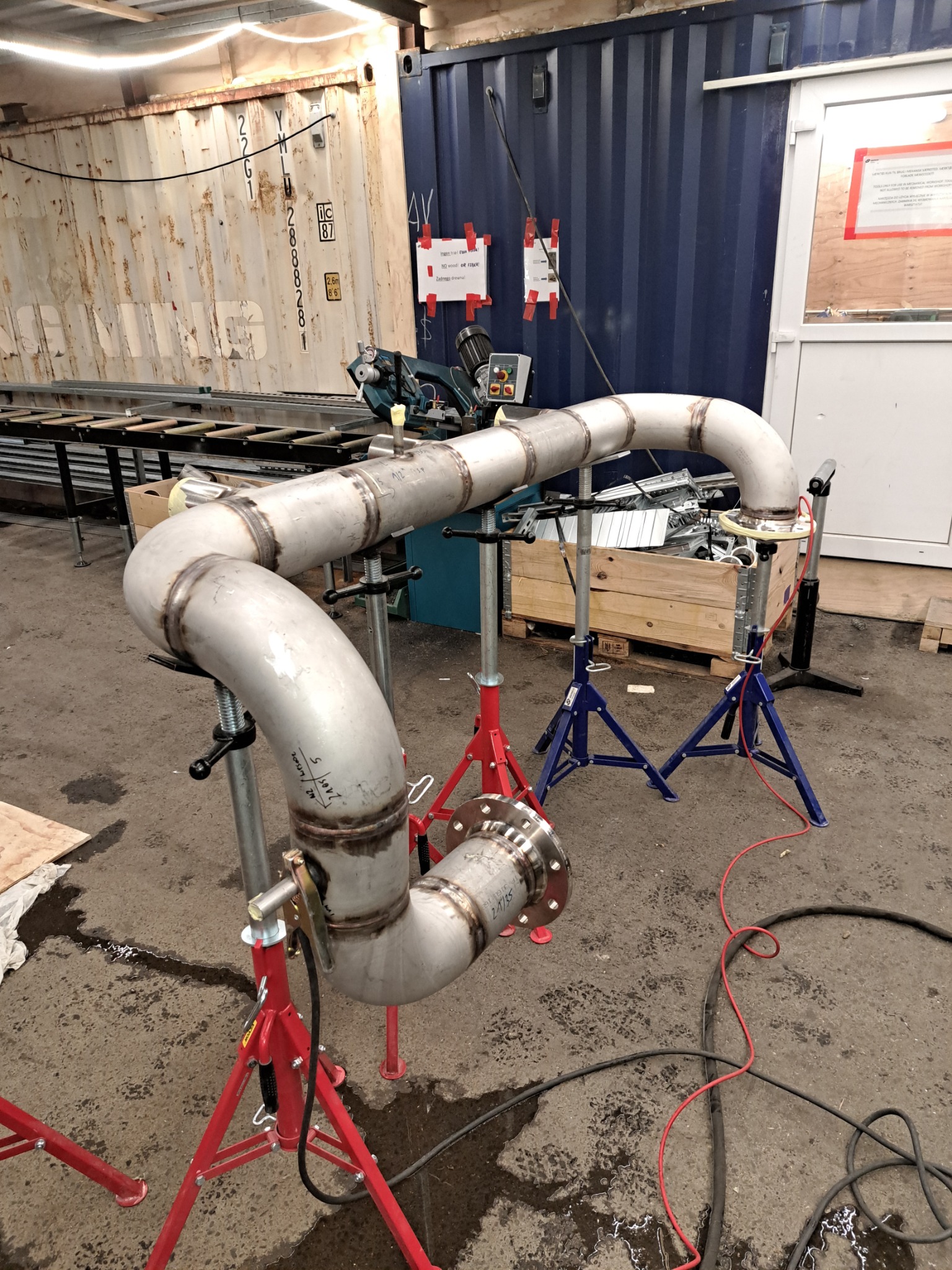 Spawany rurociąg ze stali nierdzewnej, ułożony na trójnogach spawalniczych, w warsztacie z kontenerami w tle. Widoczne ślady spawania i złącza kołnierzowe.