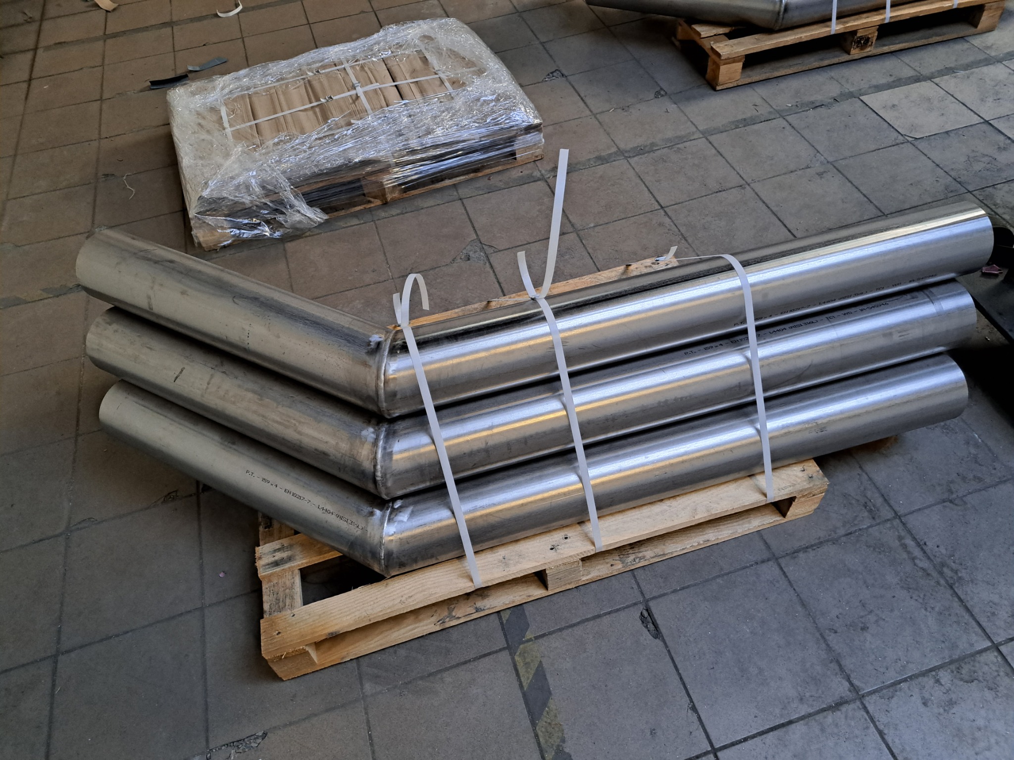 Stalowe, zespawane rury ułożone na palecie, zabezpieczone taśmą. Widoczne oznaczenia na powierzchni rur. W tle fragmenty podłogi i zapakowany element.