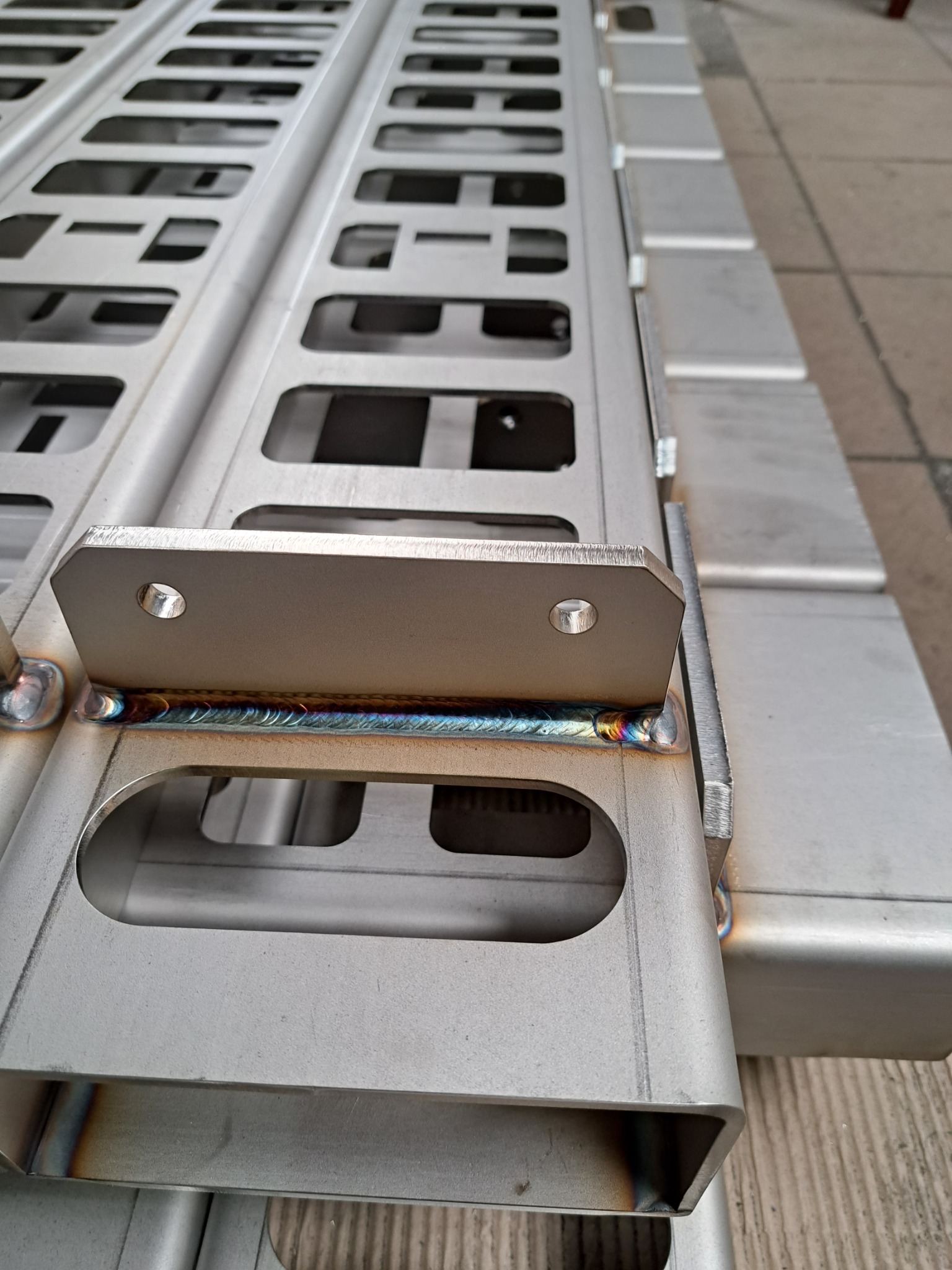 Szczegółowe zbliżenie spawu łączącego element z otworami na metalowej rampie. Widoczne tęczowe zabarwienie spoiny i regularne otwory w konstrukcji.