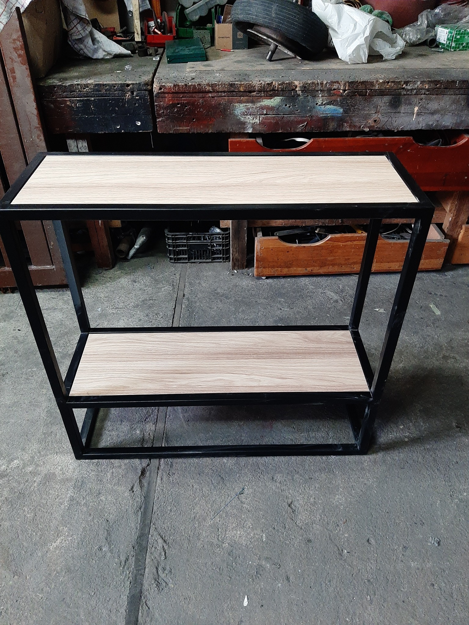 Metalowy stolik z drewnianymi półkami, czarna rama, surowe otoczenie warsztatu, widoczne ślady pracy, unikalny design.