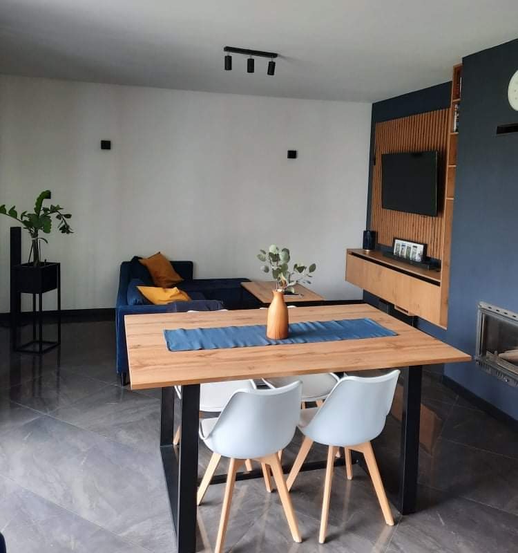 Stół z drewnianym blatem i czarnymi metalowymi nogami w nowoczesnym salonie z niebieską ścianą i elementami z drewna. Widoczne krzesła i kanapa.