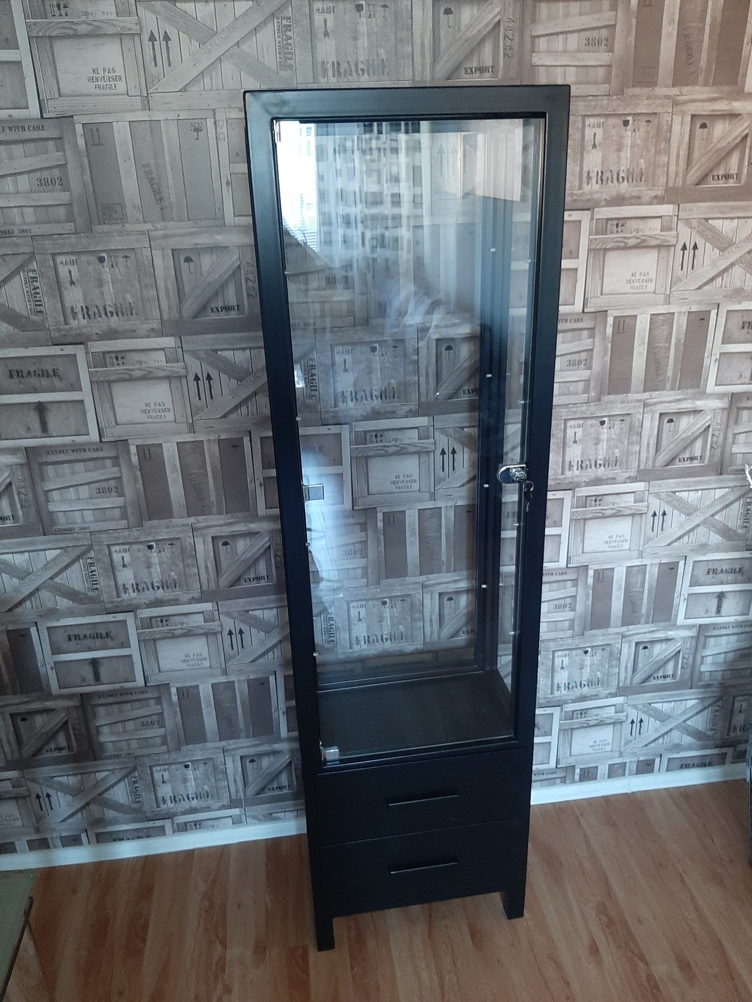 Czarna, metalowa witryna z szufladami i przeszkloną górną częścią, zamykana na klucz, na tle ściany z wzorem skrzyń transportowych.