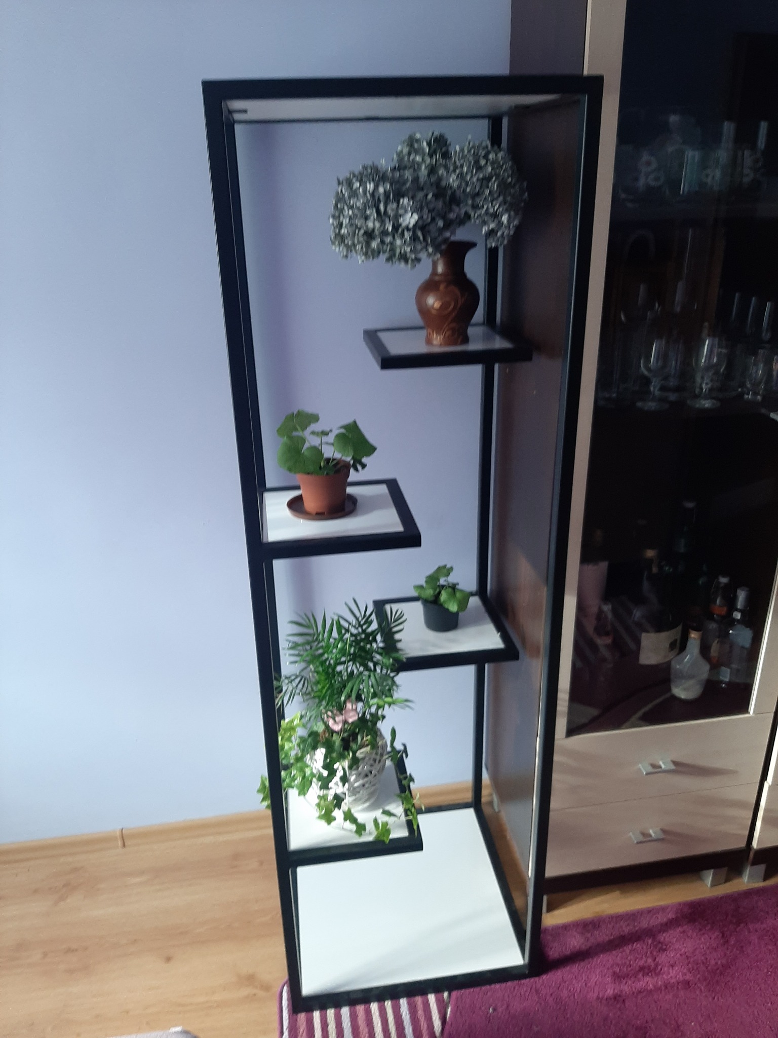 Spawana, czarna, metalowa konstrukcja regału z asymetrycznie rozmieszczonymi półkami, na których stoją rośliny doniczkowe i wazon z kwiatami.