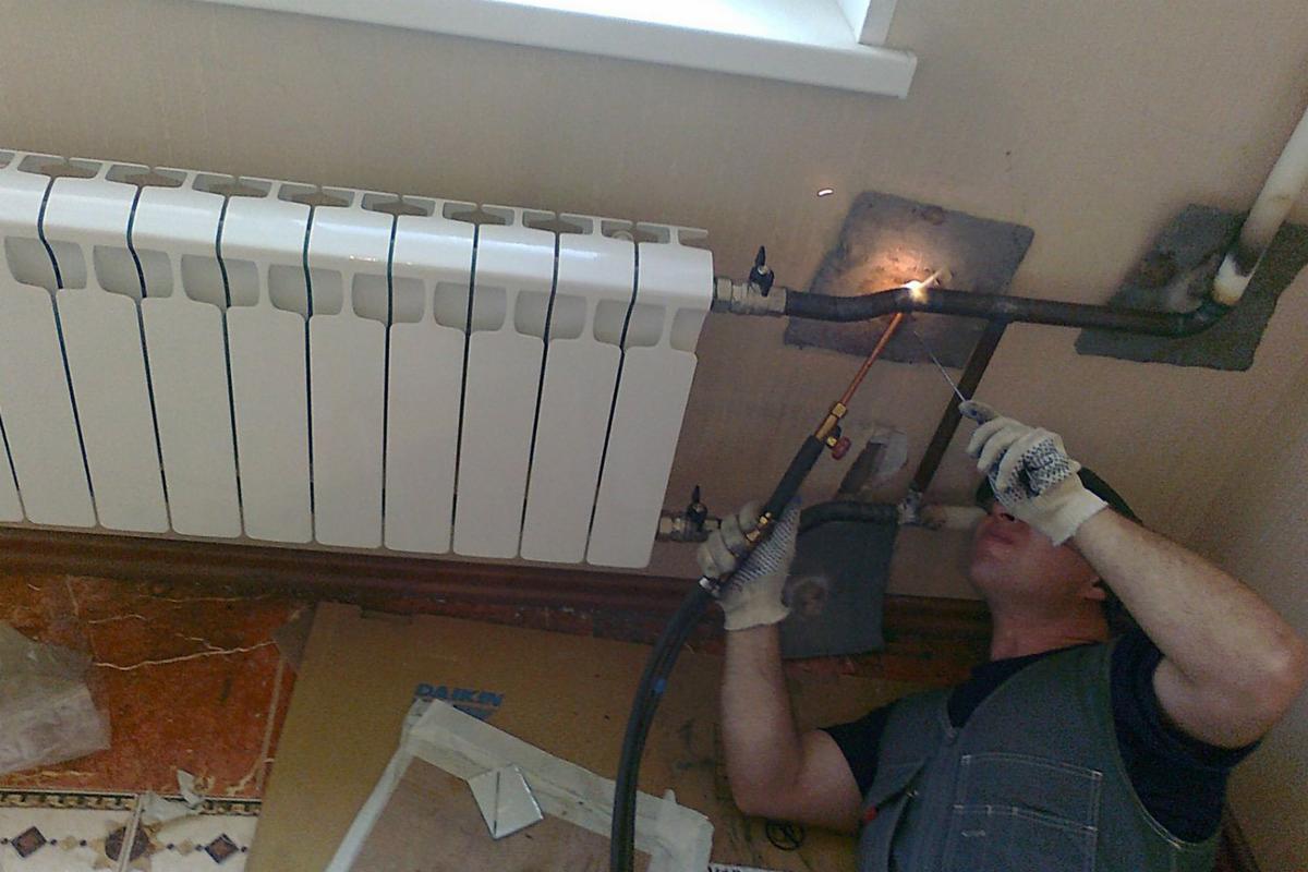 Spawanie rury instalacji centralnego ogrzewania przy grzejniku panelowym, widoczne iskry i palnik w dłoni spawacza w rękawicy ochronnej, tło stanowi ściana z widocznymi śladami prac instalacyjnych.