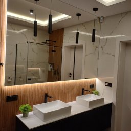 Usługi Ogólnobudowlane Marpol - Nowoczesna łazienka z dwoma kwadratowymi umywalkami nablatowymi, czarną armaturą, podświetlanym lustrem i drewnianą ścianą w tle, widoczne odbicie kabiny prysznicowej.