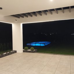 Usługi Ogólnobudowlane Marpol - Nowoczesny taras z płytek w odcieniach beżu, oświetlony listwami LED, z widokiem na podświetlony basen z zadaszeniem w ogrodzie nocą.