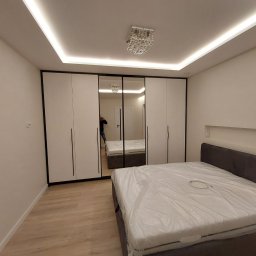 Usługi Ogólnobudowlane Marpol - Nowoczesna sypialnia z białymi szafami przesuwnymi z czarnymi ramami, lustrzanymi drzwiami, kryształowym żyrandolem i łóżkiem z szarym wezgłowiem, zabezpieczonym folią ochronną.