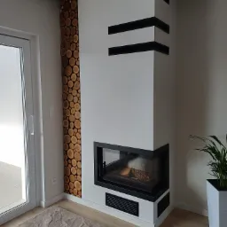 Nowoczesny kominek narożny z czarnymi akcentami i dekoracyjną ścianą z drewnianych krążków w jasnym wnętrzu z rośliną doniczkową.