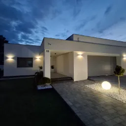 Nowoczesny dom jednorodzinny z płaskim dachem oświetlony zewnętrznymi lampami ściennymi i kulami ledowymi wzdłuż brukowanej ścieżki, numer domu 13 widoczny nad wejściem, zmierzch.