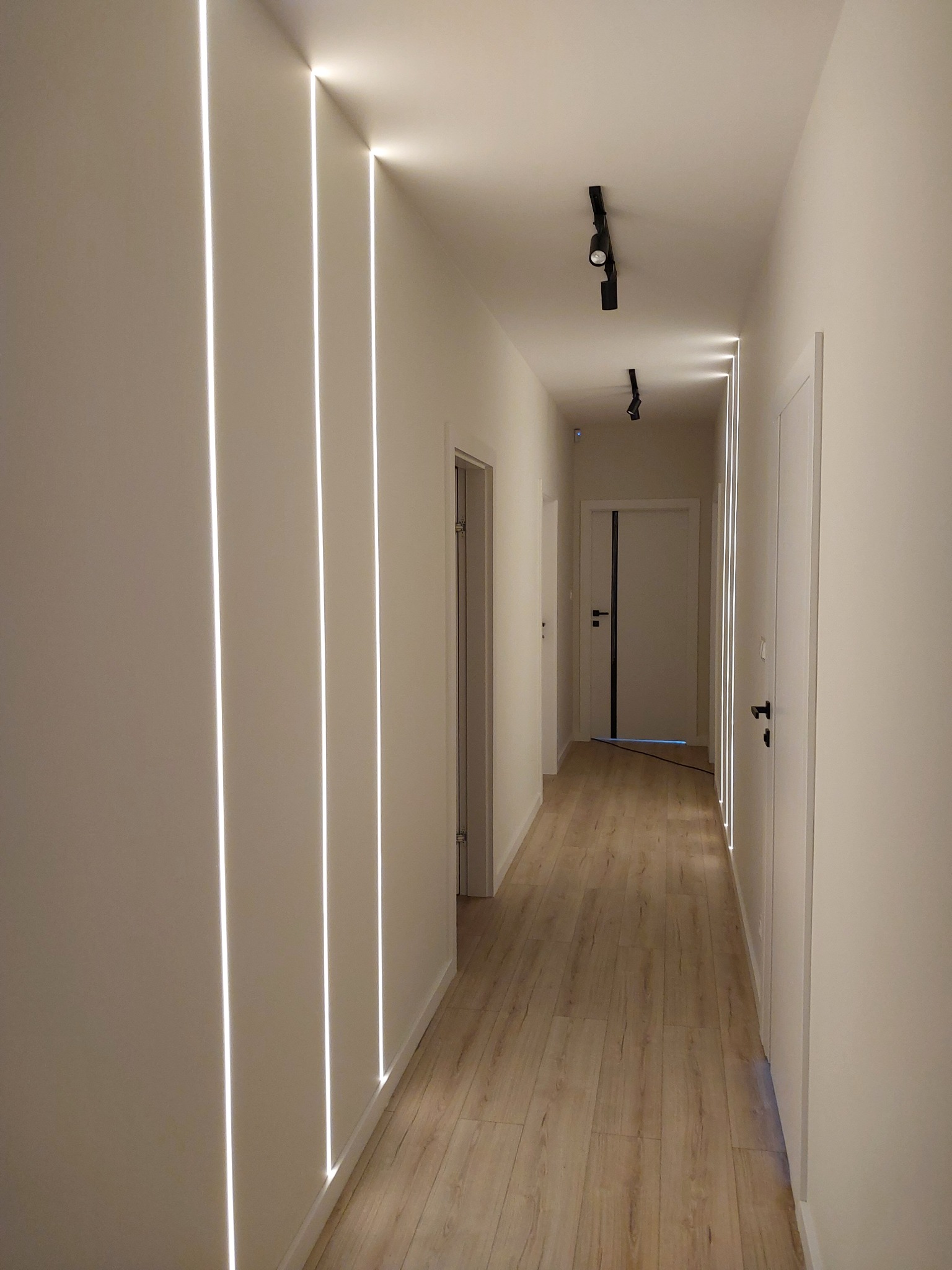 Korytarz z podświetlanymi pionowymi liniami LED w ścianach, drewnopodobna podłoga, czarne reflektory sufitowe i białe drzwi z czarnymi akcentami.
