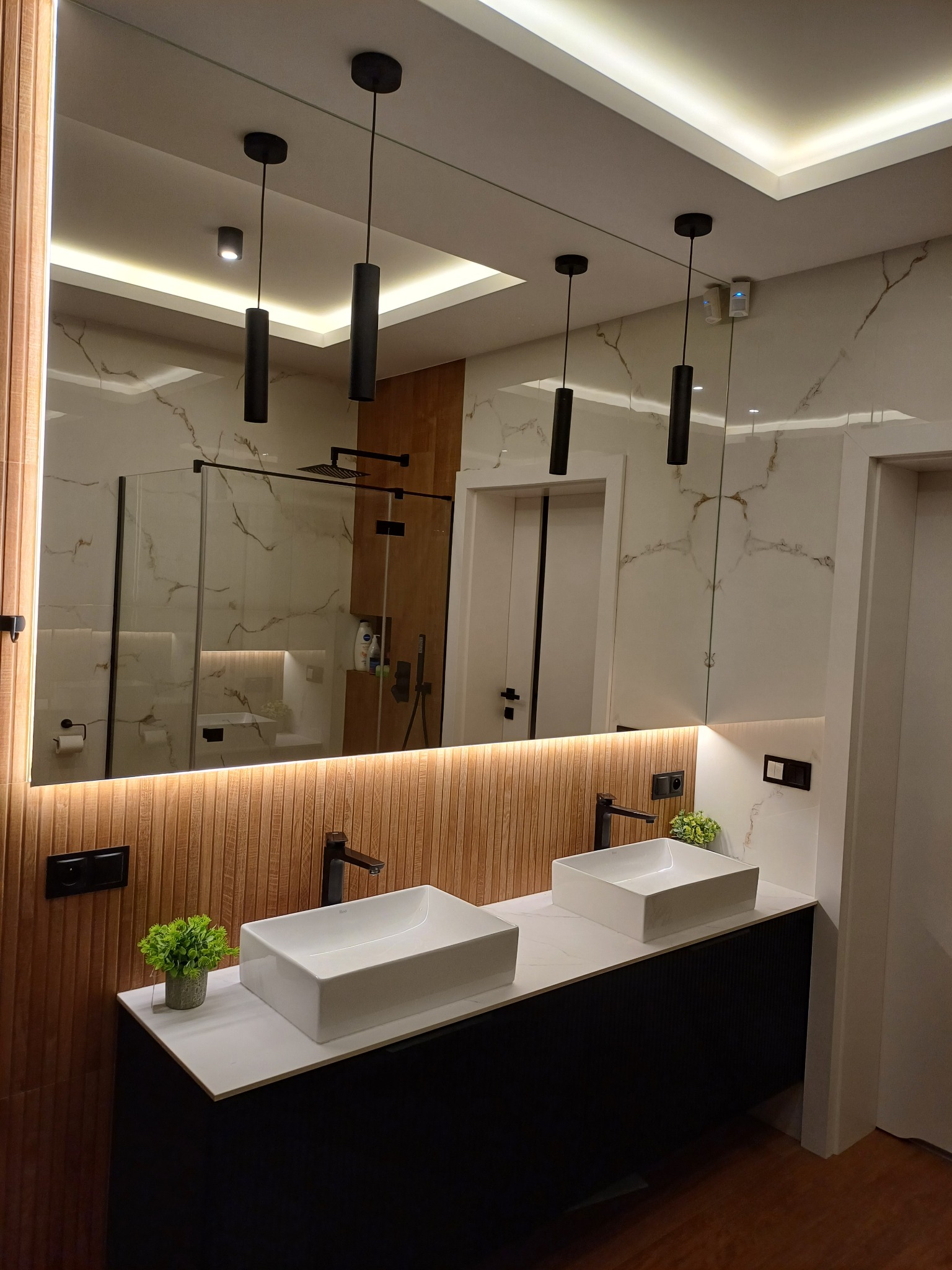 Nowoczesna łazienka z dwoma kwadratowymi umywalkami nablatowymi, czarną armaturą, podświetlanym lustrem i drewnianą ścianą w tle, widoczne odbicie kabiny prysznicowej.