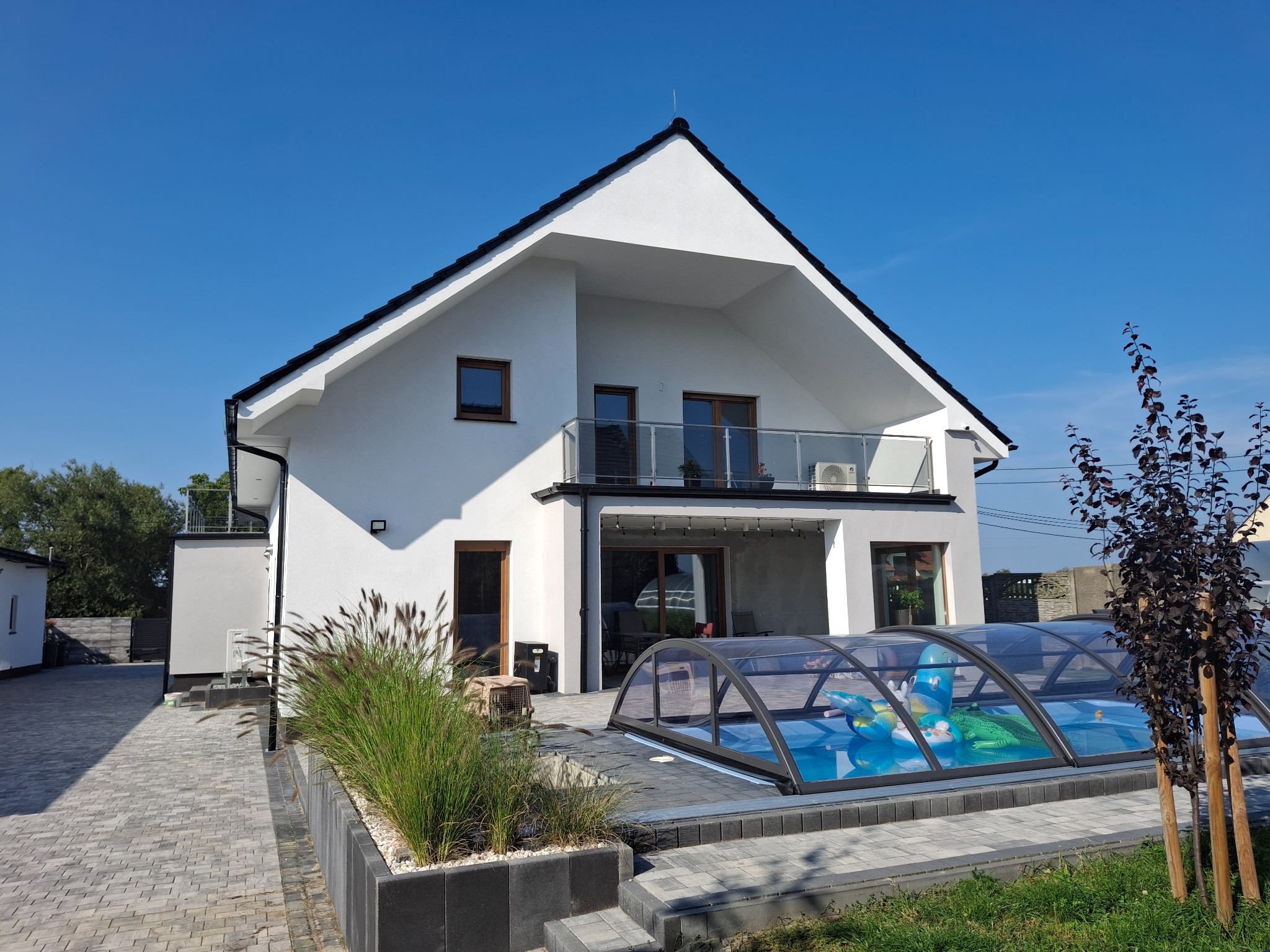 Nowoczesny, dwukondygnacyjny dom z białymi ścianami i czarnym dachem, z zadaszonym basenem w ogrodzie, widok z zewnątrz w słoneczny dzień.