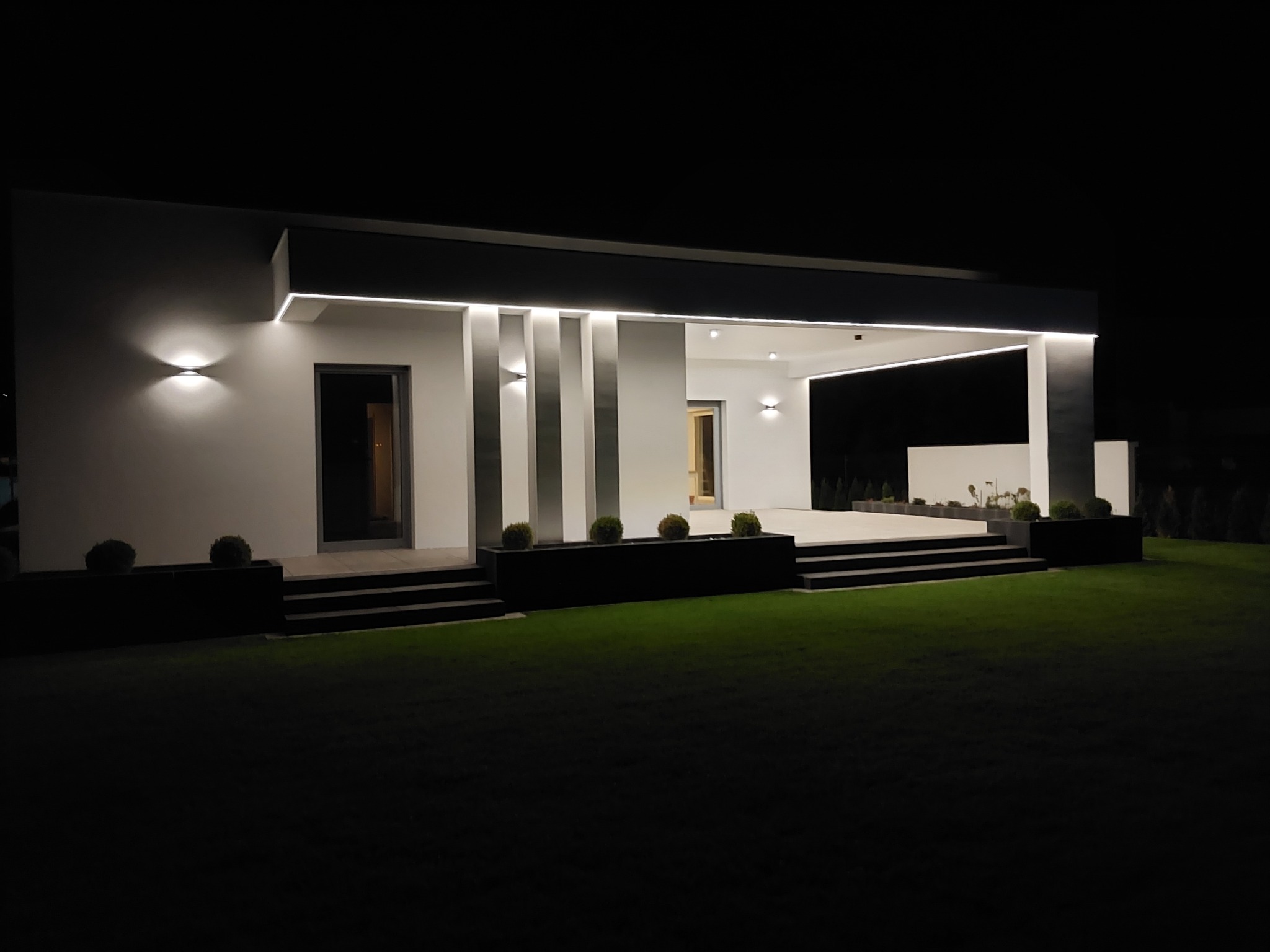 Nowoczesny dom jednorodzinny z płaskim dachem oświetlony nocą, z minimalistyczną elewacją w kolorze białym i szarymi kolumnami, z podświetleniem LED podkreślającym architekturę i zadbanym trawnikiem.