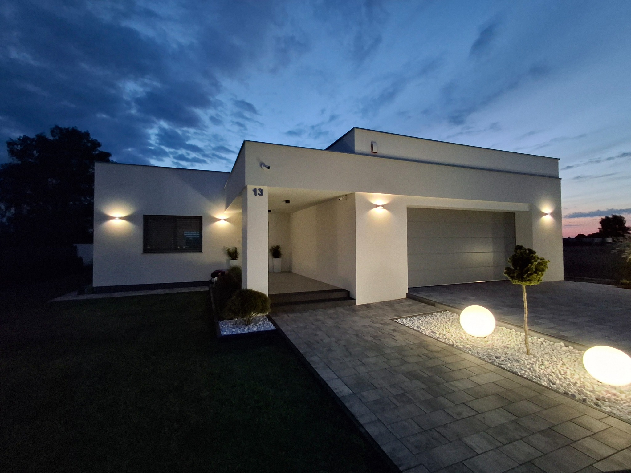 Nowoczesny, parterowy dom z płaskim dachem oświetlony po zmierzchu, z numerem 13 nad wejściem i kulistymi lampami wzdłuż ścieżki.
