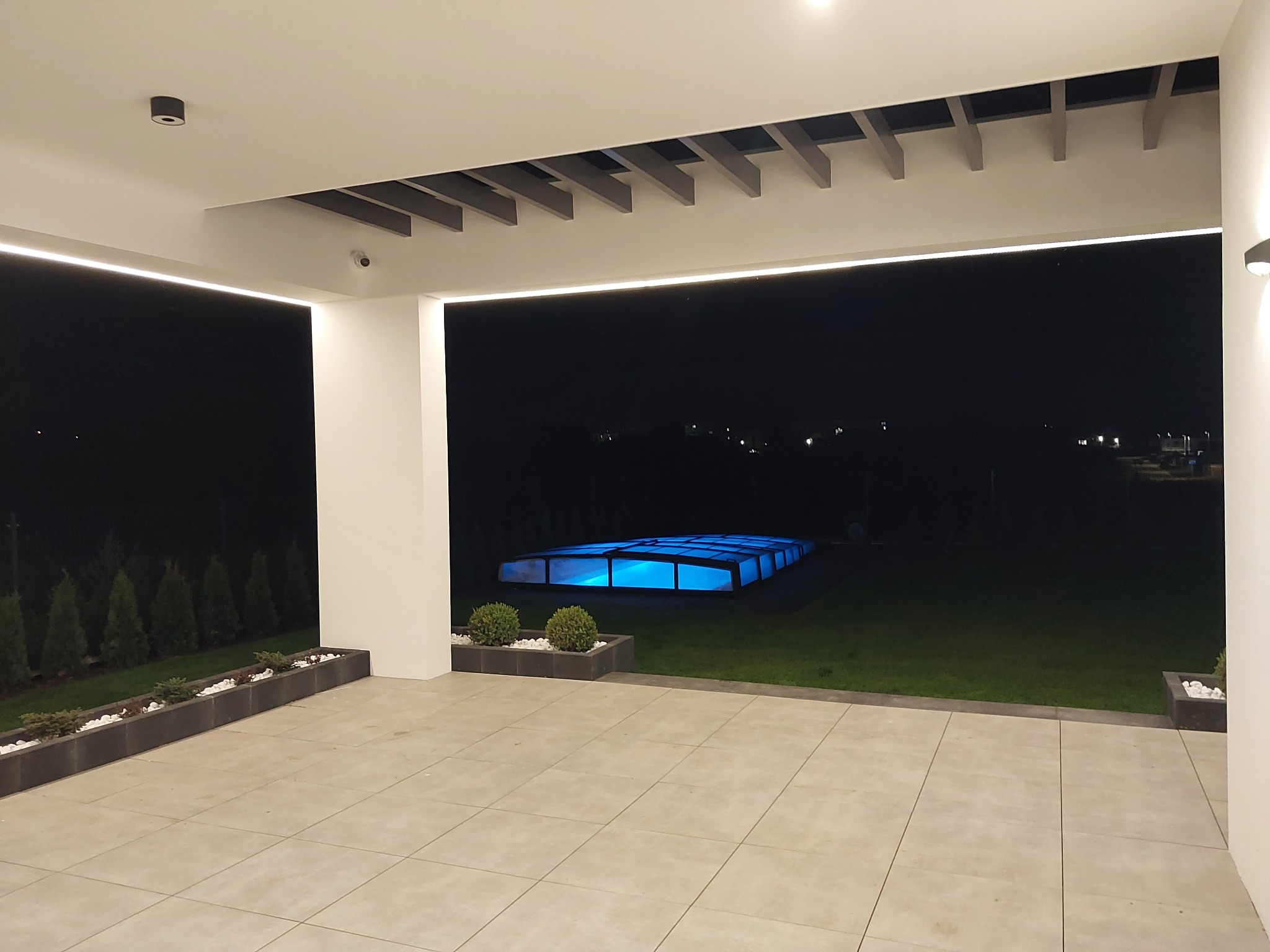 Nowoczesny taras z płytek w odcieniach beżu, oświetlony listwami LED, z widokiem na podświetlony basen z zadaszeniem w ogrodzie nocą.