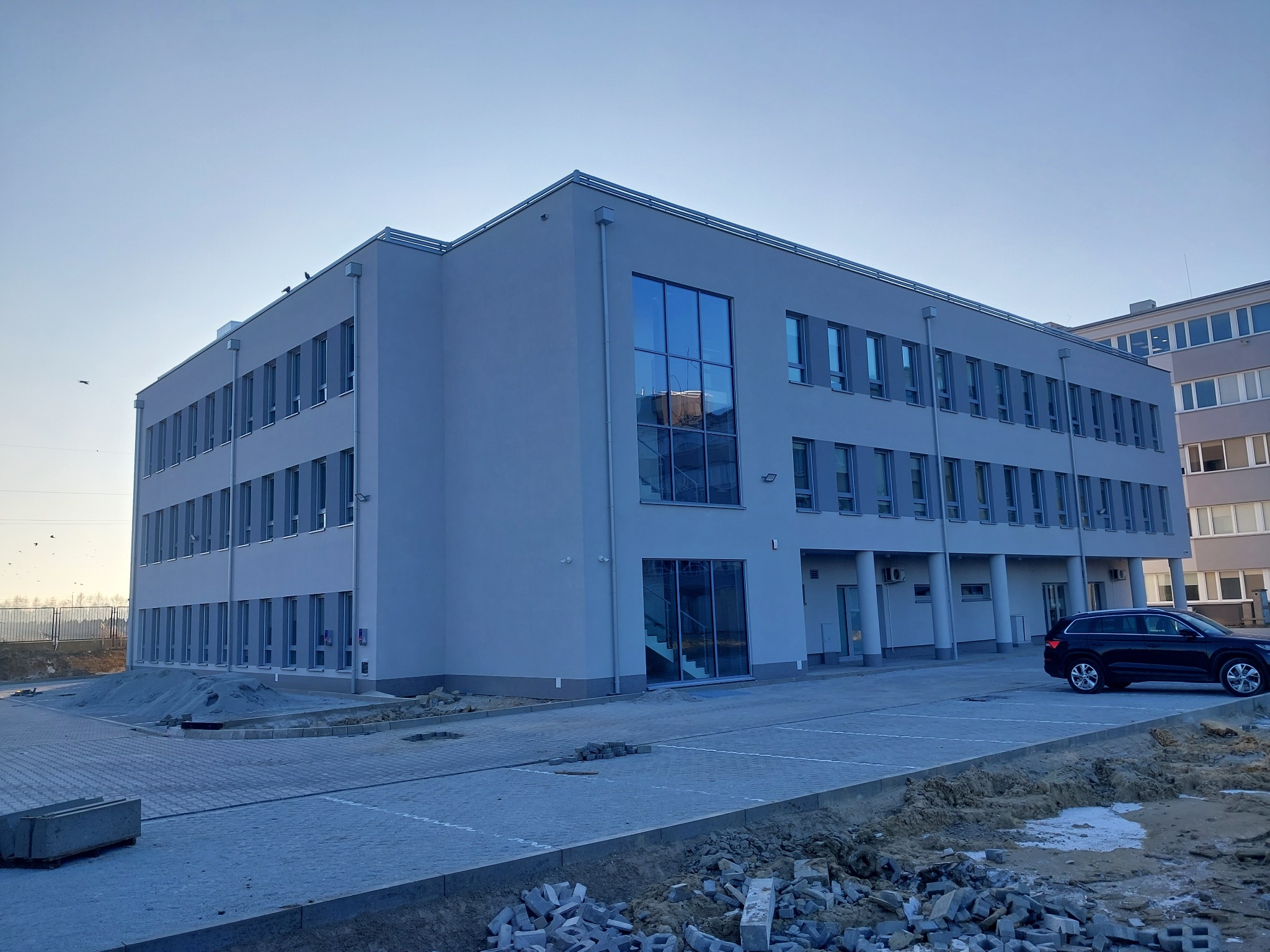 Nowoczesny, trzypiętrowy budynek biurowy w odcieniach szarości z dużymi oknami, częściowo wykończony, z czarnym samochodem osobowym zaparkowanym obok i z kostką brukową na placu przed budynkiem.