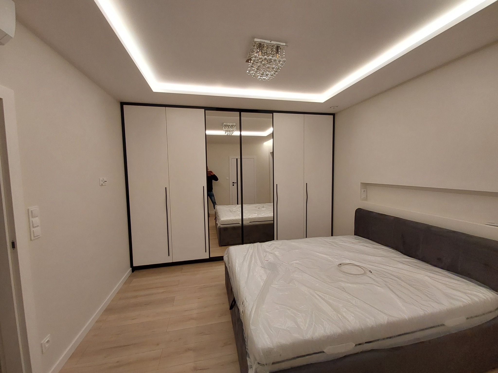 Nowoczesna sypialnia z białymi szafami przesuwnymi z czarnymi ramami, lustrzanymi drzwiami, kryształowym żyrandolem i łóżkiem z szarym wezgłowiem, zabezpieczonym folią ochronną.
