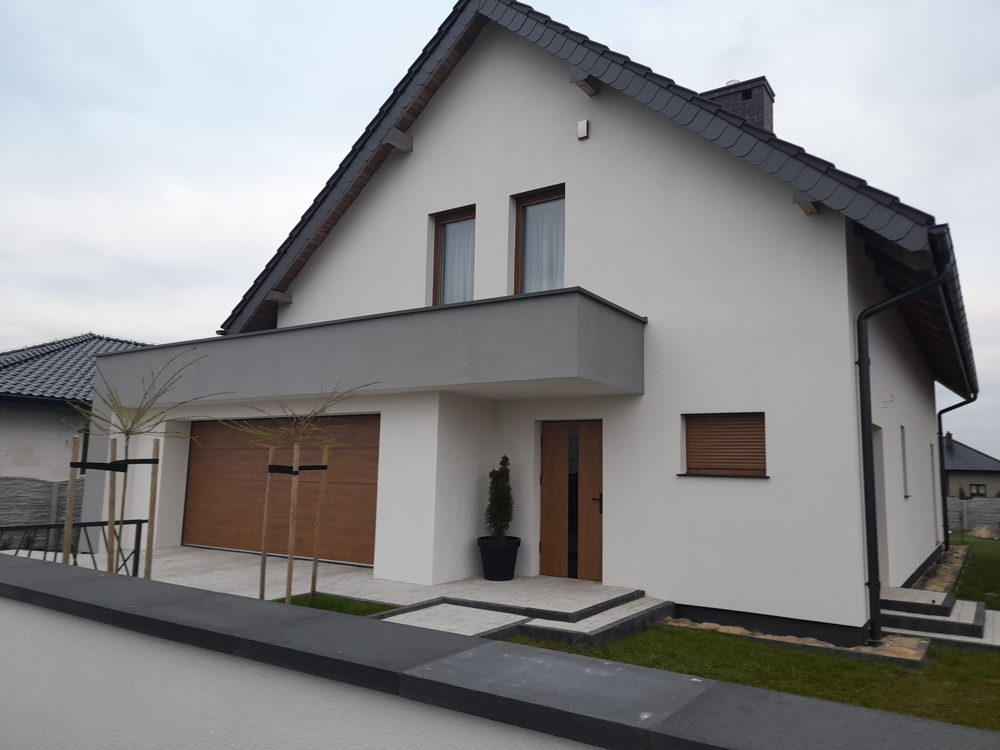 Nowoczesny dom jednorodzinny z białymi ścianami, grafitowym dachem z dachówki karpiówki, drewnianą bramą garażową i drzwiami wejściowymi, z małym balkonem i dwoma młodymi drzewkami przy garażu.