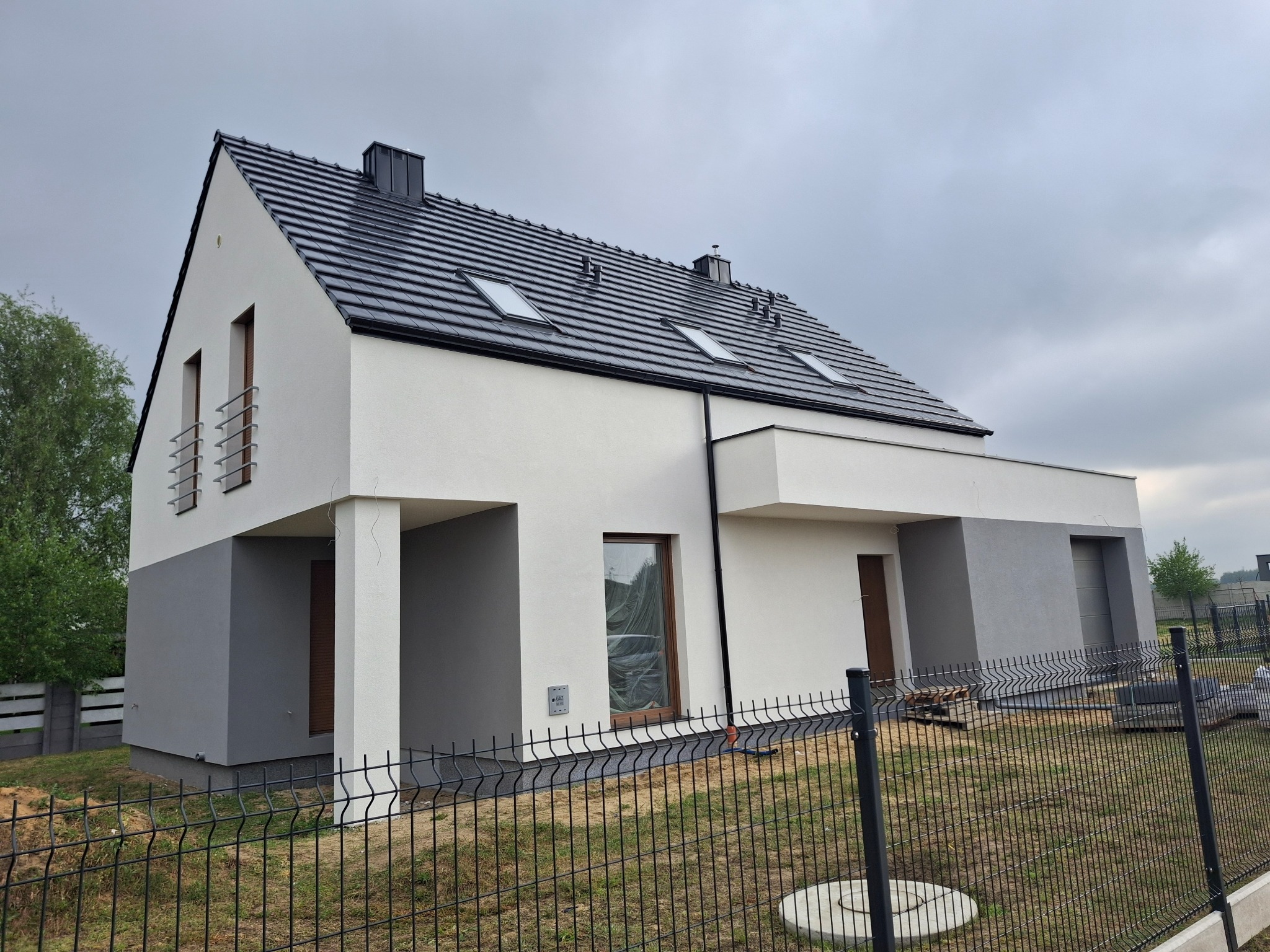 Nowoczesny dom murowany w Ostrowie Wielkopolskim z dwukolorową elewacją, czarną dachówką i widocznym ogrodzeniem panelowym. Okna zabezpieczone folią malarską.