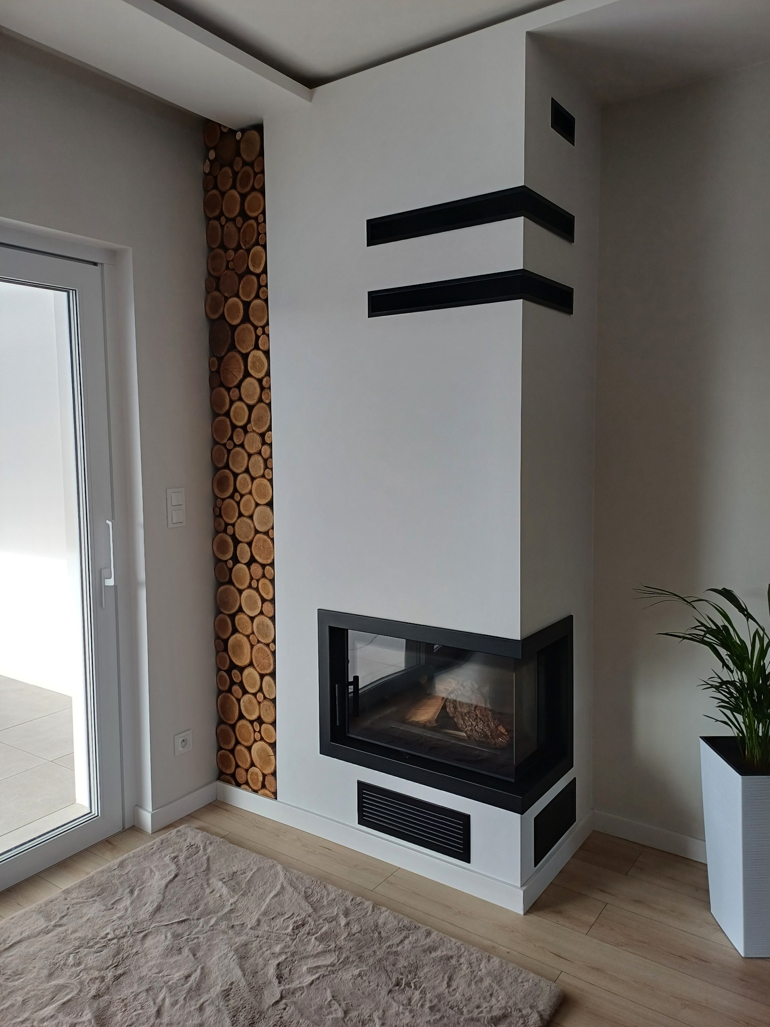 Nowoczesny kominek narożny z czarnymi akcentami i dekoracyjną ścianą z drewnianych krążków w jasnym wnętrzu z rośliną doniczkową.