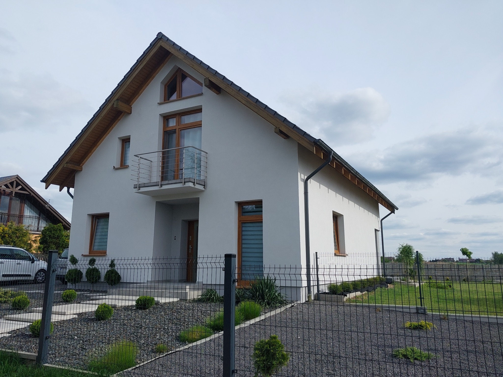 Nowoczesny dom jednorodzinny z białymi ścianami, drewnianymi oknami i balkonem ze stali nierdzewnej, otoczony czarnym, metalowym ogrodzeniem i ozdobnym ogrodem z kamyków i niskich krzewów.