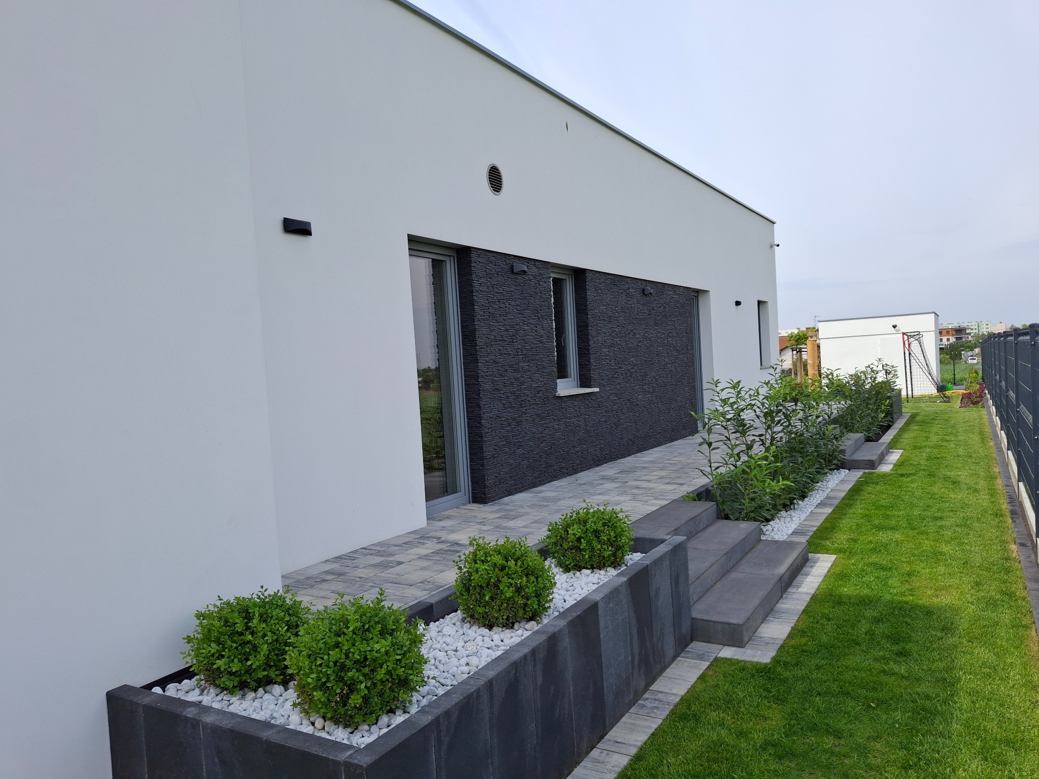 Nowoczesny dom z białymi ścianami, ciemną elewacją z kamienia łupanego, szarą kostką brukową i zielonym trawnikiem z ozdobnymi krzewami w betonowych donicach wypełnionych białymi kamieniami.