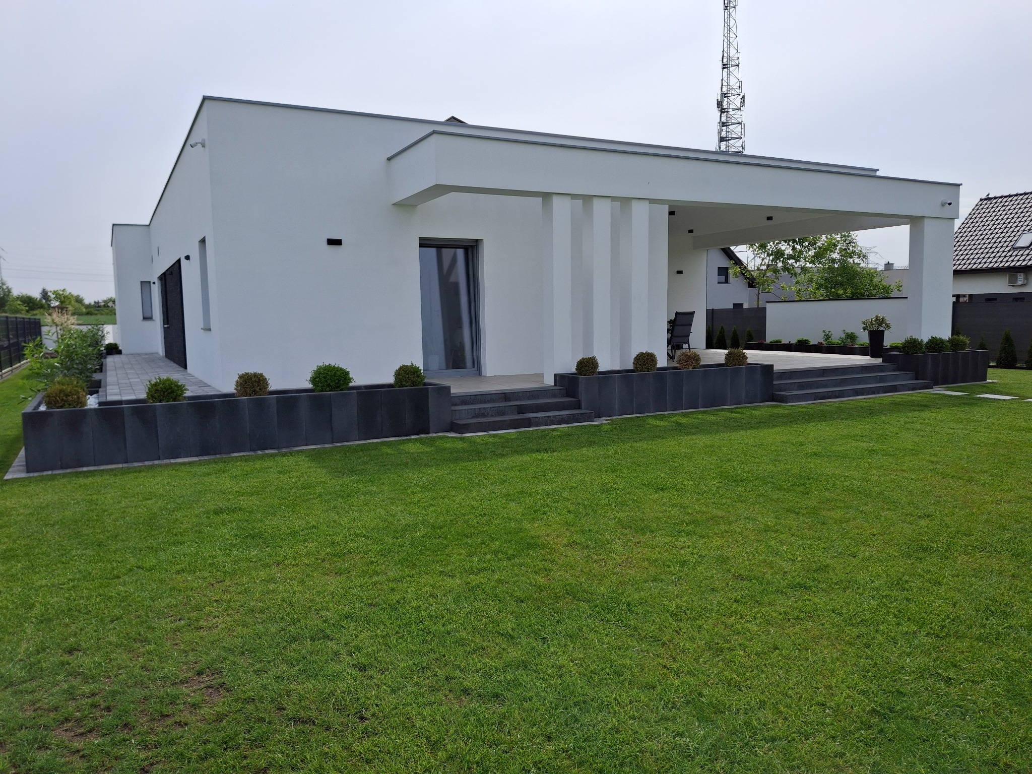 Nowoczesny, parterowy dom z białymi ścianami i zadaszonym tarasem, otoczony zadbanym trawnikiem i ciemnymi donicami z kulistymi krzewami.