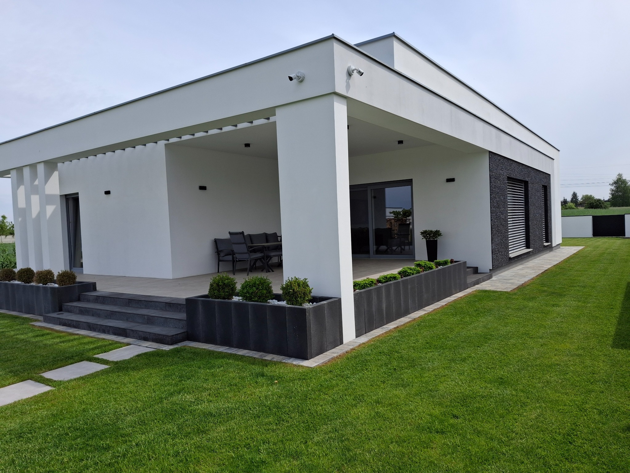 Nowoczesny dom jednorodzinny z białymi ścianami i czarnymi elementami dekoracyjnymi, widok z zewnątrz na taras z meblami ogrodowymi, schodami i donicami z roślinami, kamery monitoringu na elewacji.