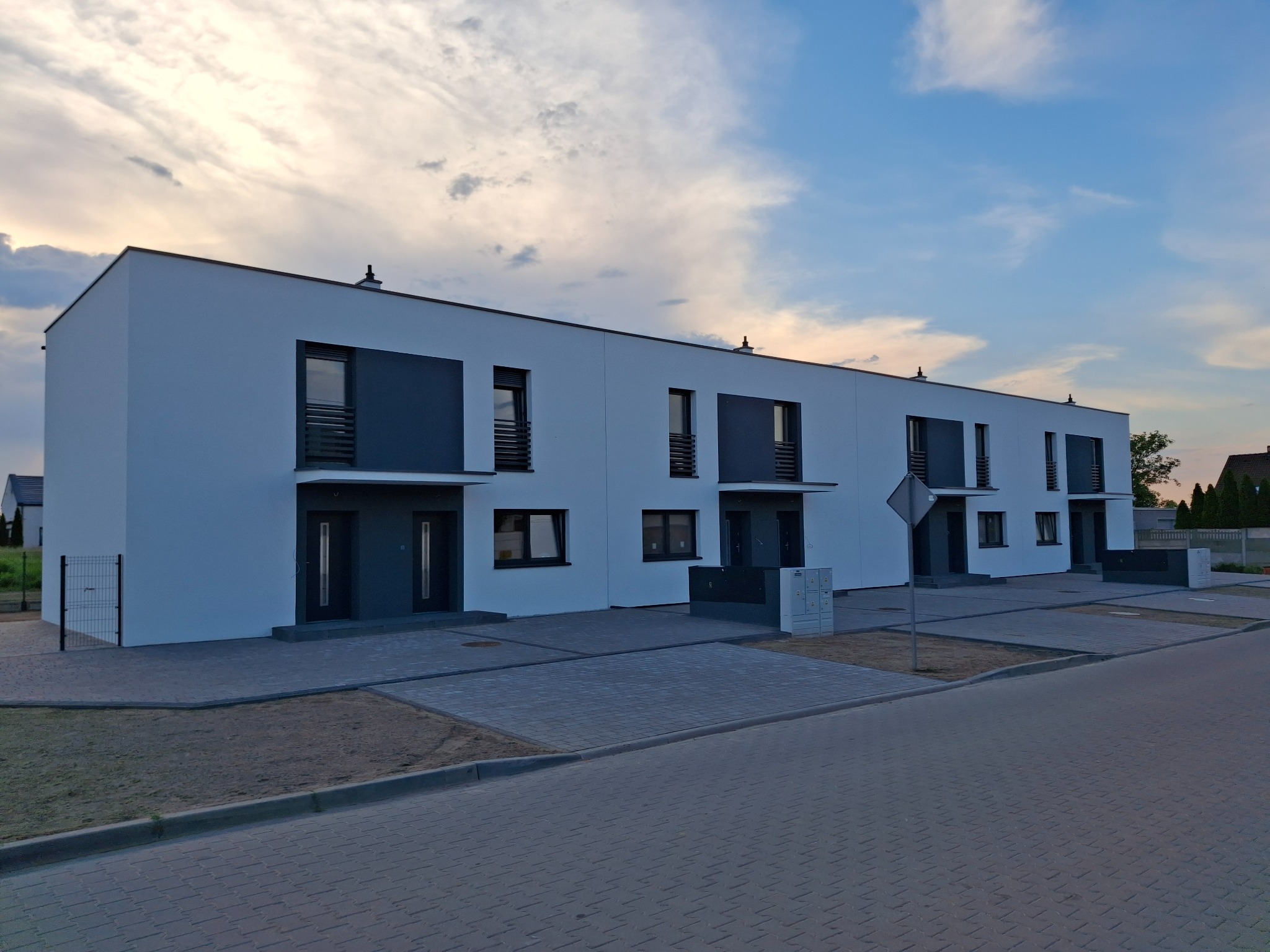 Szereg identycznych, nowoczesnych domów murowanych z płaskimi dachami i antracytowymi akcentami, widok z poziomu ulicy na tle zachmurzonego nieba.