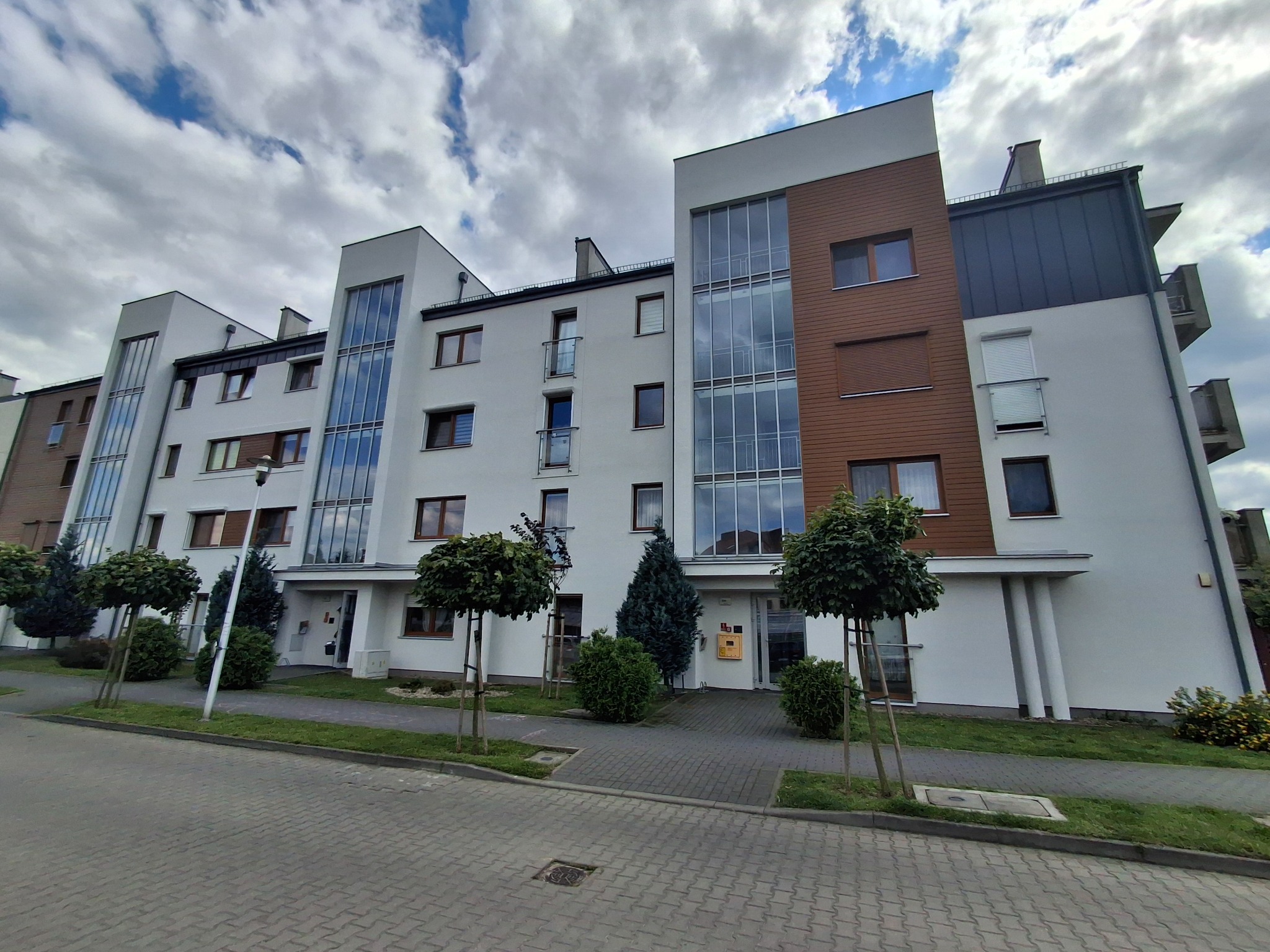 Nowoczesny, trzypiętrowy budynek mieszkalny z białymi ścianami, brązowymi panelami elewacyjnymi i dużymi oknami, otoczony niskimi drzewami i krzewami na tle pochmurnego nieba.