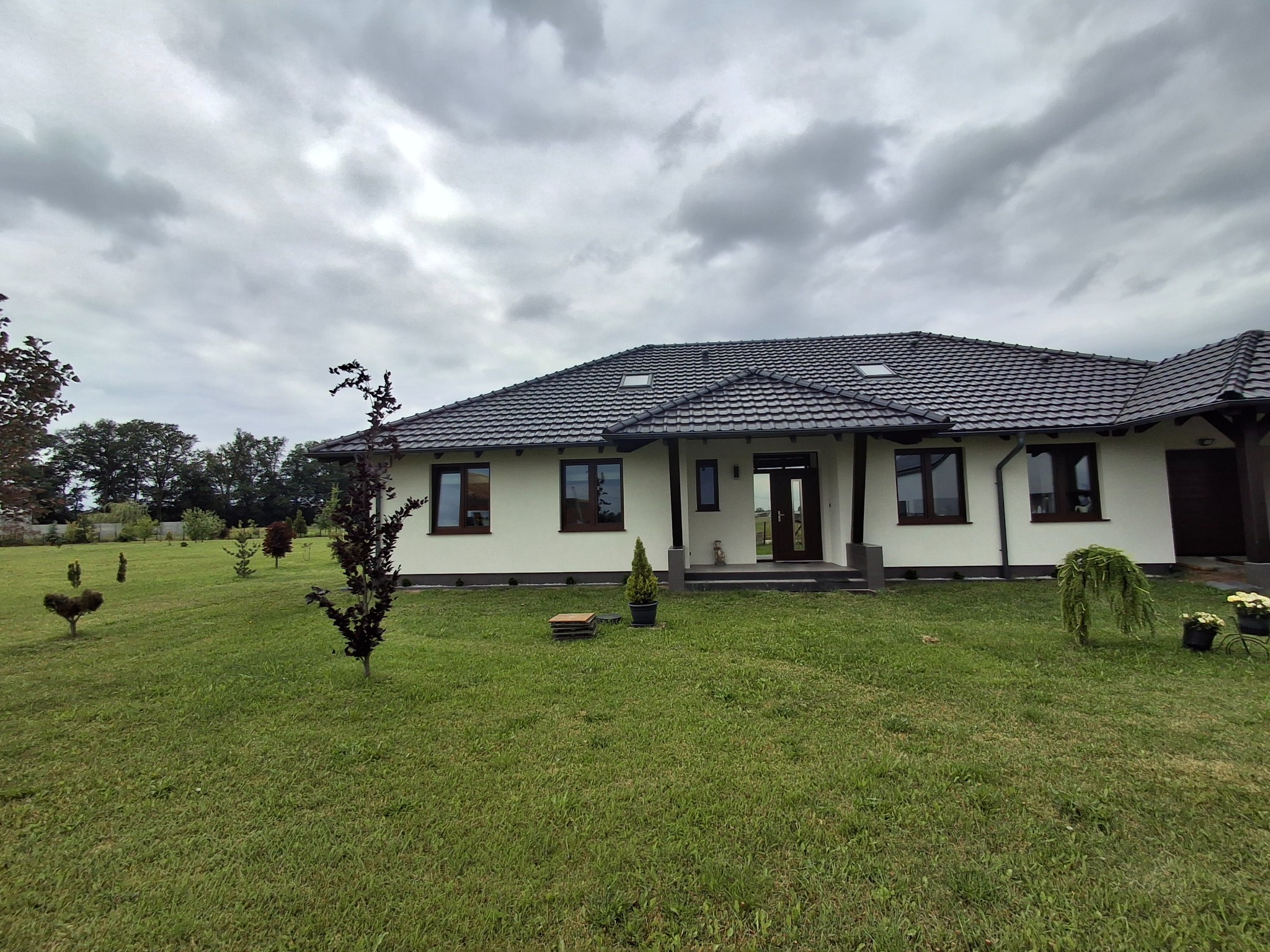 Parterowy dom z białymi ścianami i ciemną dachówką, widok z ogrodu w pochmurny dzień. Na trawniku ozdobne krzewy i drzewka.