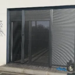 Zewnętrzny widok domu z przesuwną moskitierą plisowaną w kolorze antracytowym, zamontowaną na wejściu, obok rolety zewnętrznej. Na pierwszym planie rośliny w kwadratowych donicach i niebieskie klapki.