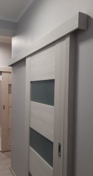 Białe drzwi przesuwne wewnętrzne z szarymi wstawkami szklanymi, widoczne w pozycji częściowo otwartej, z zamontowanym systemem przesuwnym. Widok z boku, zarys futryny w tle.