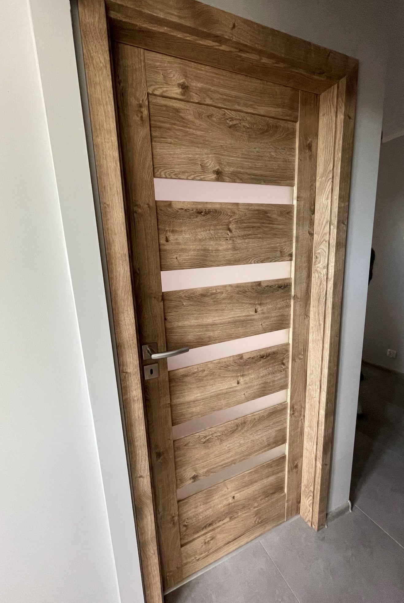 Drewniane drzwi wewnętrzne z czterema poziomymi przeszkleniami, widoczne od strony klamki, zamontowane w białej ścianie.