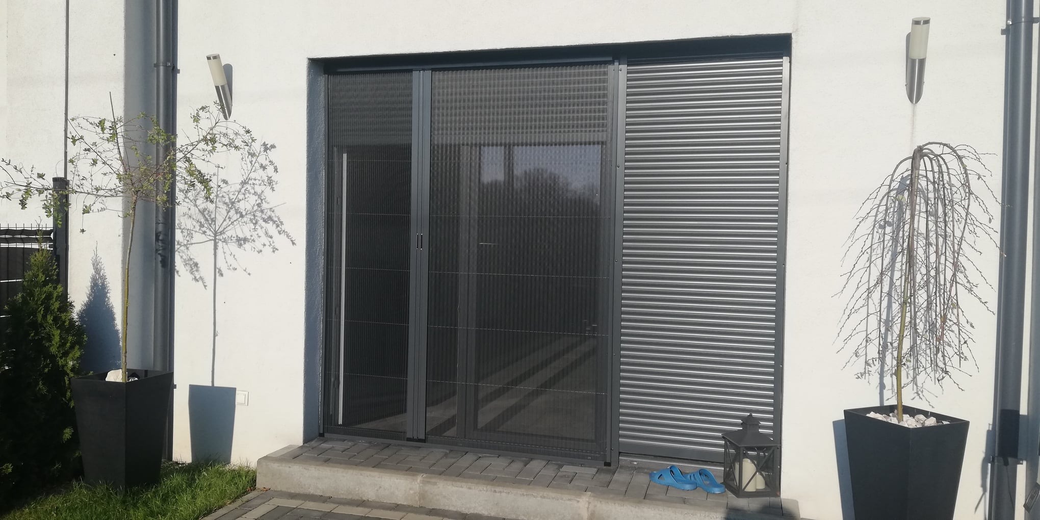 Zewnętrzny widok domu z przesuwną moskitierą plisowaną w kolorze antracytowym, zamontowaną na wejściu, obok rolety zewnętrznej. Na pierwszym planie rośliny w kwadratowych donicach i niebieskie klapki.