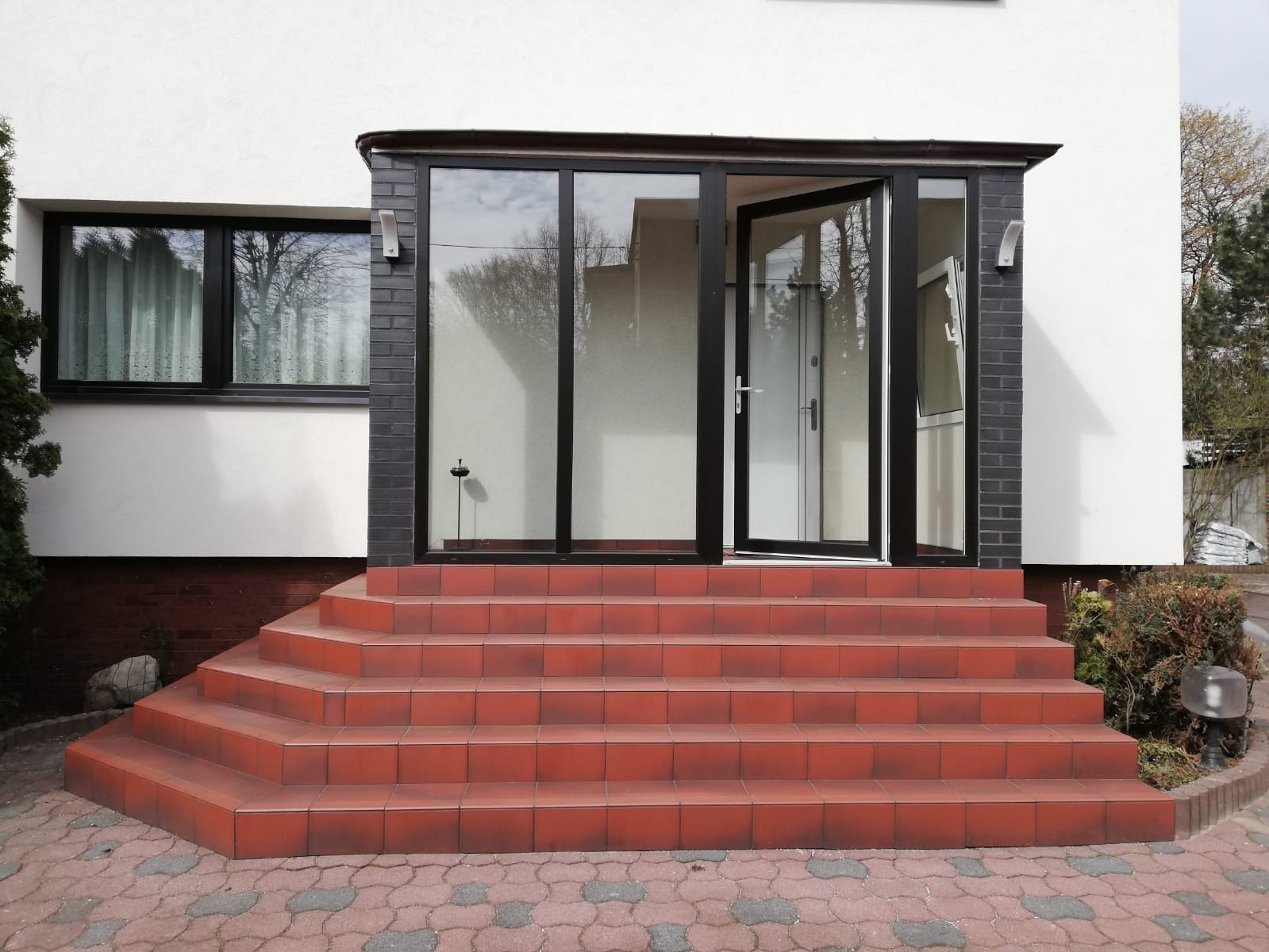 Zadaszone wejście do domu z otwartymi drzwiami balkonowymi w czarnej ramie, widoczne schody z czerwonej terakoty i okno z zasłonami.
