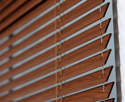 Zamknięte żaluzje drewniane w odcieniu orzecha, widoczne sznurki i metalowe zaczepy, delikatne światło wpadające przez szpary.