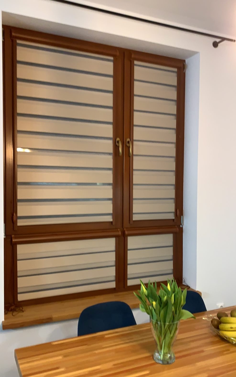 Drewniane okno z zamontowanymi roletami dzień-noc, widoczne z perspektywy wnętrza, z bukietem tulipanów w wazonie na stole w pierwszym planie.