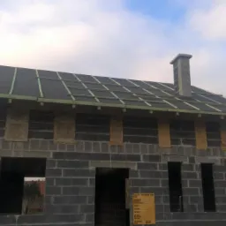 Surowy stan domu z szarej cegły z fragmentem dachu pokrytego papą. Widoczne drewniane łaty i komin. Tablica informacyjna na ścianie. Stan surowy otwarty.