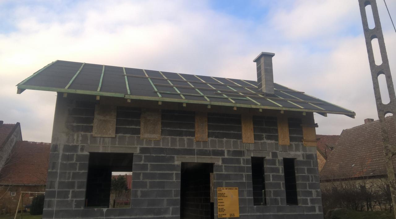 Surowy stan domu z szarej cegły z fragmentem dachu pokrytego papą. Widoczne drewniane łaty i komin. Tablica informacyjna na ścianie. Stan surowy otwarty.