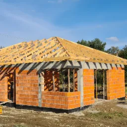 Budowa domu jednorodzinnego z czerwonej cegły, widoczne ściany i drewniana konstrukcja dachu w trakcie montażu, teren budowy z tabliczką ostrzegawczą.