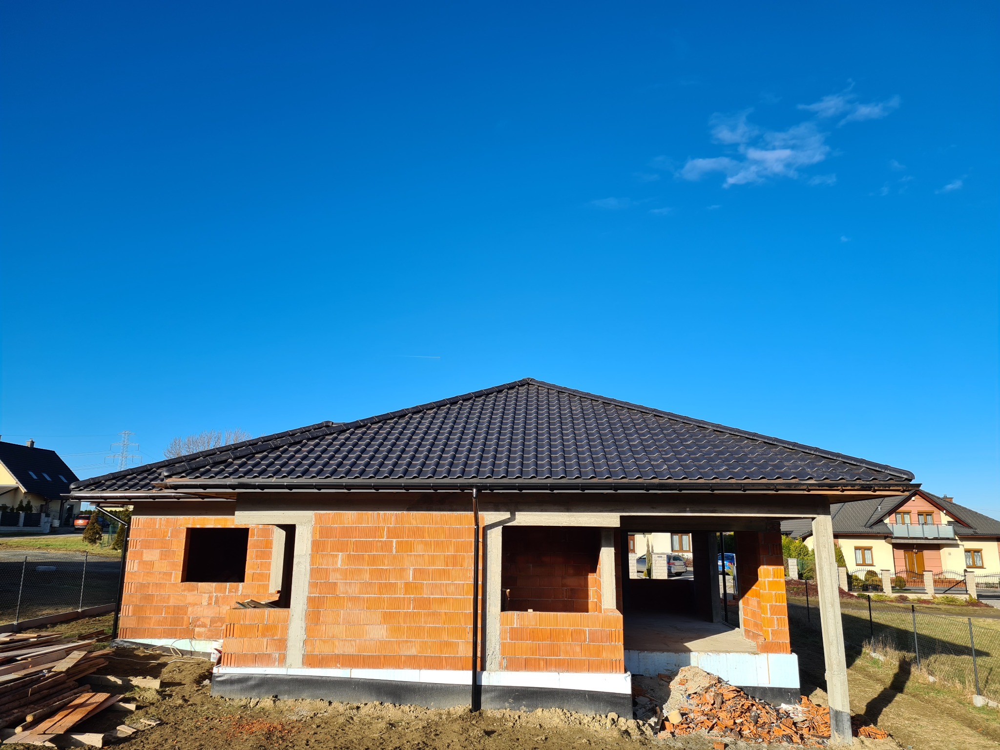 Niedokończony dom jednorodzinny z czerwonej cegły z czarnym dachem na tle błękitnego nieba, widoczne otwory okienne i drzwiowe, oraz stos cegieł przed budynkiem.