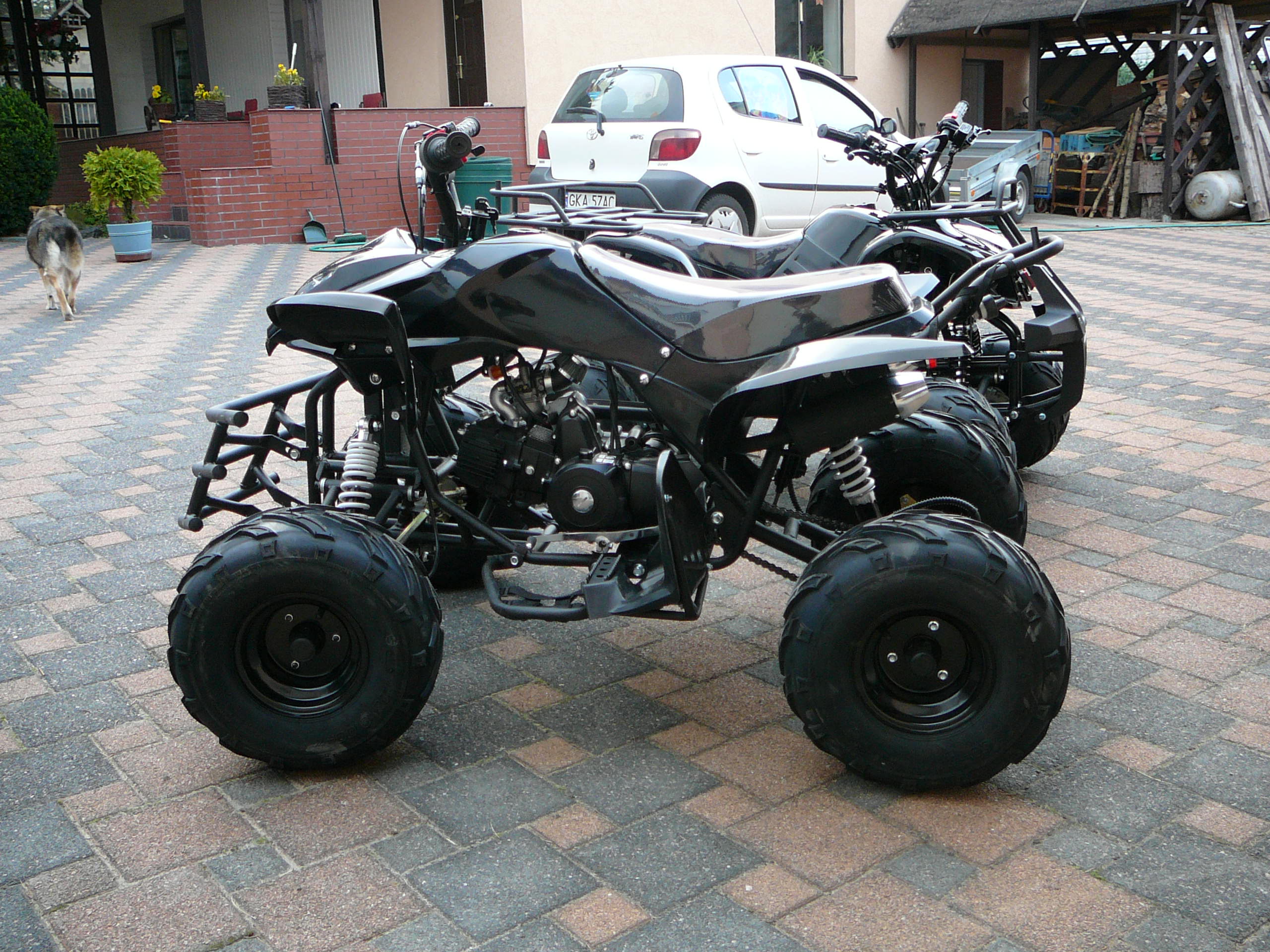 Dwa czarne quady zaparkowane na kostce brukowej w Przyjaźni, z psem w tle i budynkiem mieszkalnym.