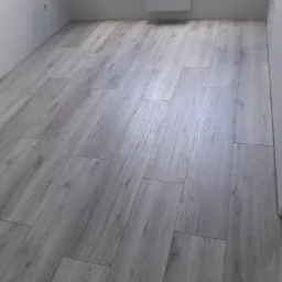 Szare panele podłogowe ułożone w jasnym pokoju z grzejnikiem i gładkimi ścianami.