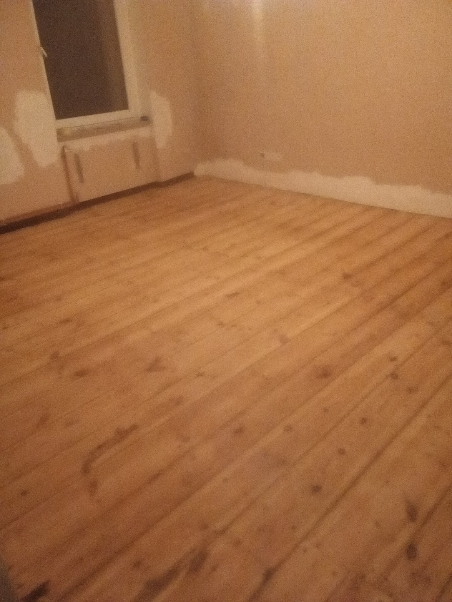 Wnętrze pokoju z ułożoną, lakierowaną podłogą z parkietu, ściany przygotowane do malowania, widoczne ślady szpachlowania przy oknie i listwach przypodłogowych.