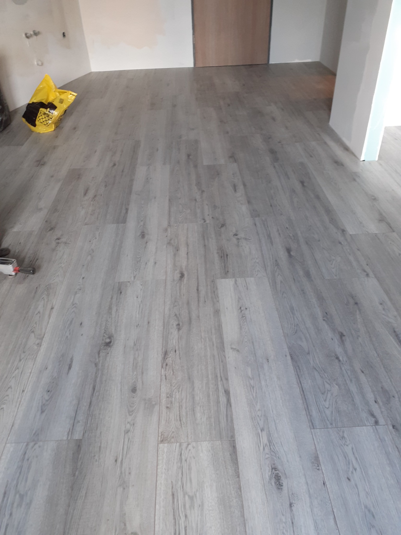 Ułożone panele podłogowe w odcieniach szarości, widoczny worek z resztkami i narzędzie montażowe w rogu pomieszczenia.